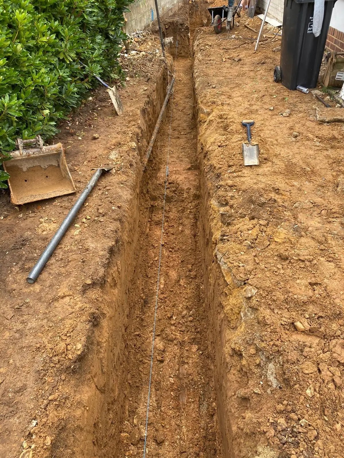 A service trench dug to a line beside the bungalow, with the digger bucket and spade alongside