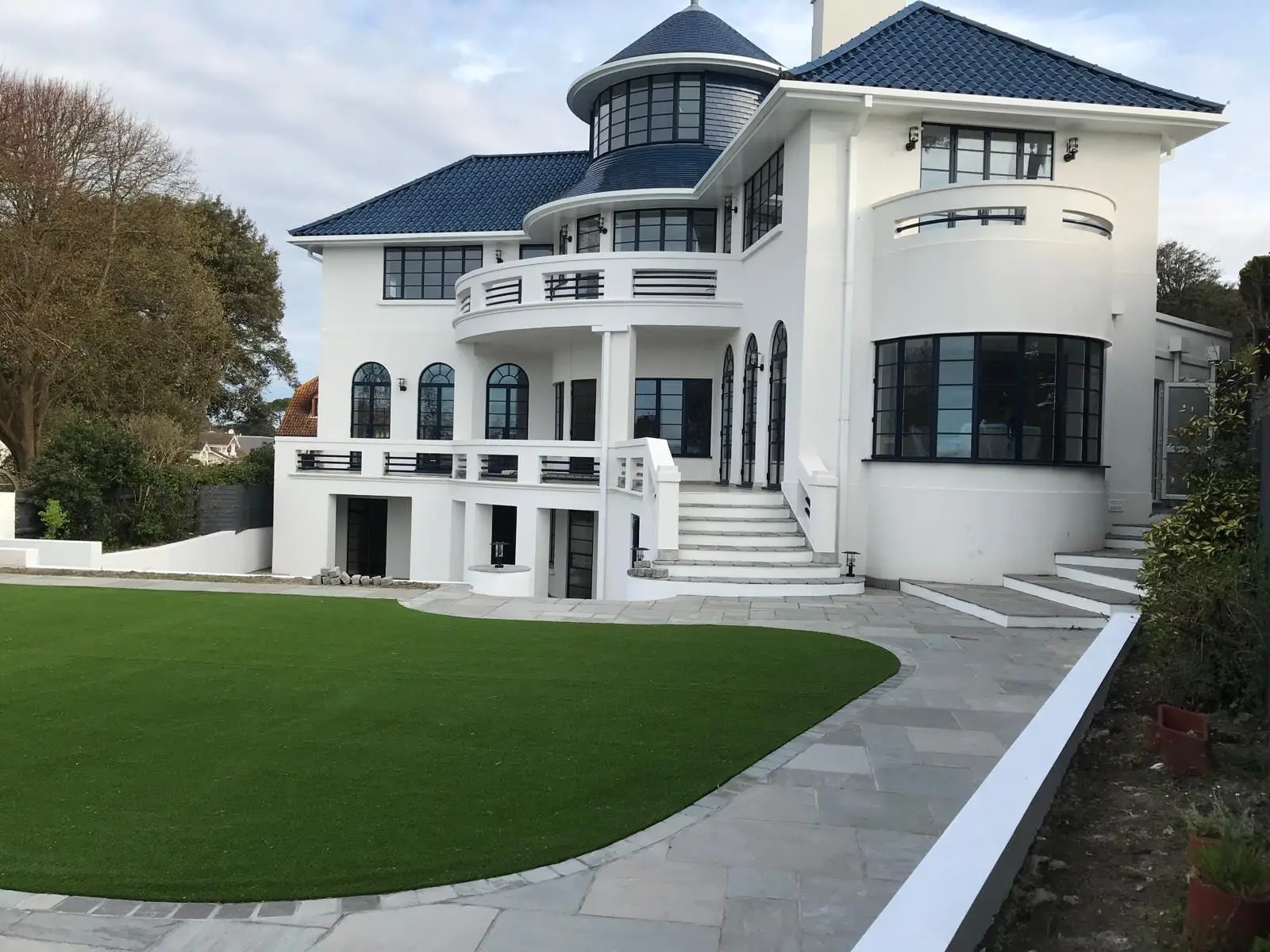 The restored Art Deco house: white render, curved balconies, arched steel windows and a blue glazed pantile roof with a turret