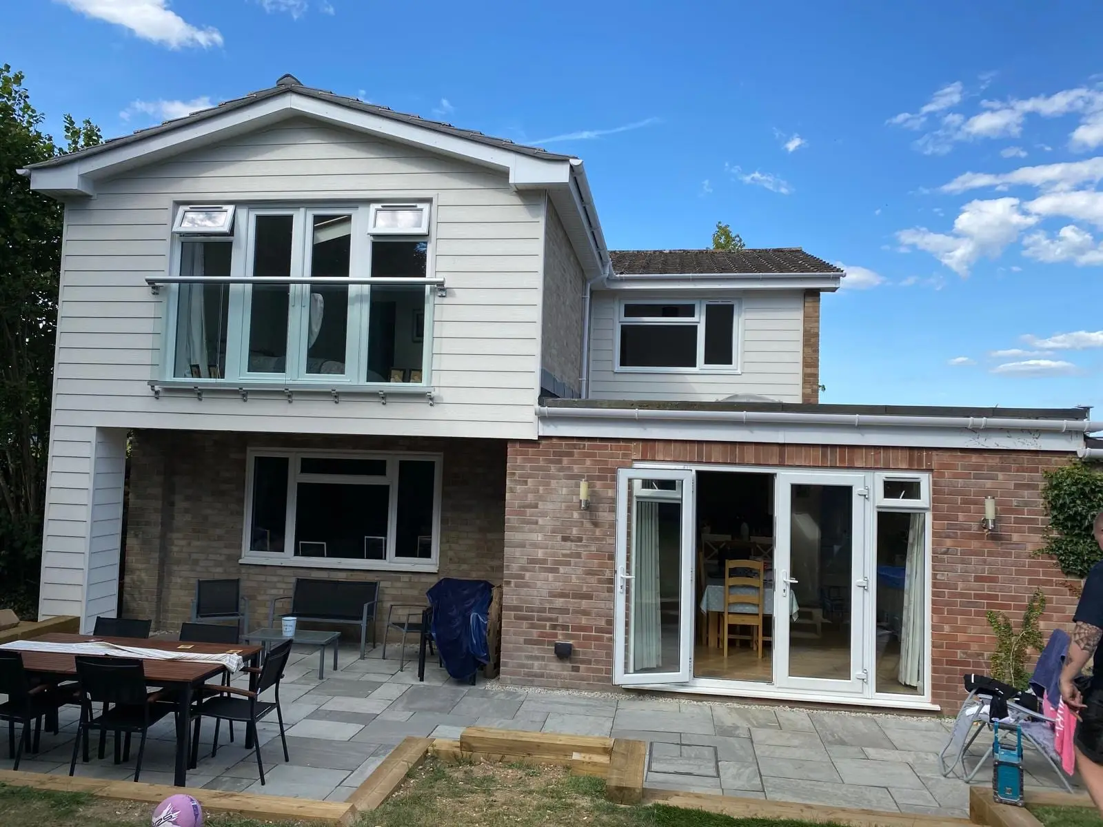 The finished rear elevation: a weatherboarded first floor extension with a glass Juliet balcony over brickwork, and a sandstone patio